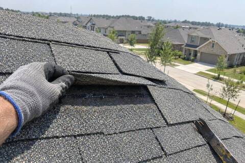 Gloved hand lifting a creased shingle tab near the ridge of a gray shingle roof, rows of newer houses beyond