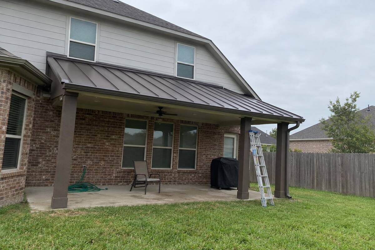 Dark bronze standing-seam metal roof over a backyard patio cover on a brick and siding house