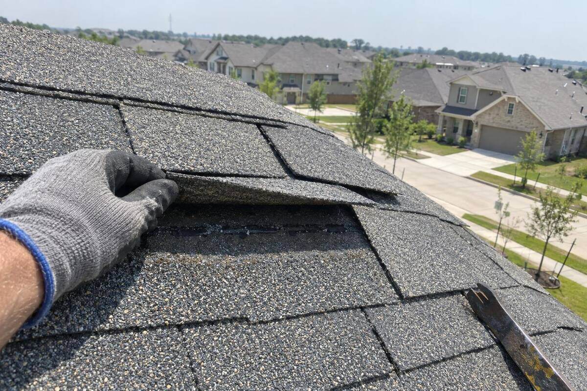 Gloved hand lifting a creased shingle tab near the ridge of a gray shingle roof, rows of newer houses beyond